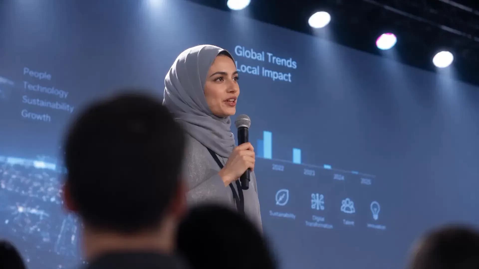 A woman in a grey hijab speaking into a microphone in front of a large screen headed “Global Trends, Local Impact” with a bar chart.